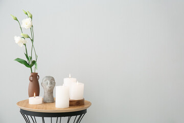 Burning candles, vase with eustoma flowers and decorative head on end table near grey wall