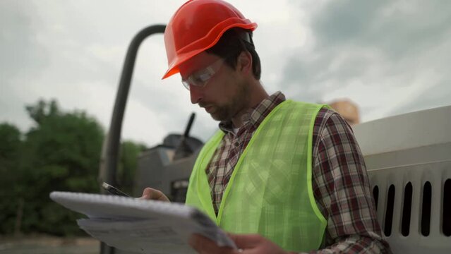Engineer builder hard hat standing against paver at construction site, checking plan of project with technical documentation and using smartphone. Worker in front of roller on building site. 