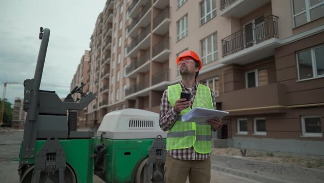 Engineer builder hard hat standing against paver at construction site, checking plan of project with technical documentation and using smartphone. Worker in front of roller on building site. 