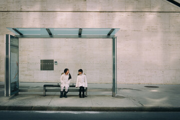 Two asian girls dressed in winter clothe sitting at a bus stop in the city