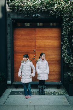 Two Young Asian Girls Standing In Front Of A Modern House Entrance Door