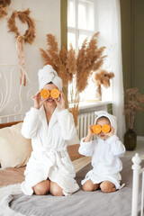 Full body faceless portrait of beautiful mother and cute little daughter having fun, sitting on bed with fresh fruit orange for face mask having spa day toghether at home - obrazy, fototapety, plakaty
