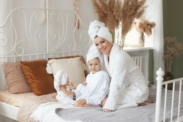 Obraz na płótnie Canvas Loving mother take care to little daughter in white bathrobes and with towel on heads after a shower playing at morning on bed at cozy home