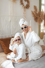 Happy mothers day concept. A little girl with her beautiful mother self-care after a shower, sitting in white bathrobes, sunglasses and with a towel on her head in a cozy bedroom at home