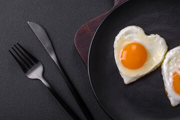 Two heart-shaped fried eggs on a black ceramic plate on a dark concrete background