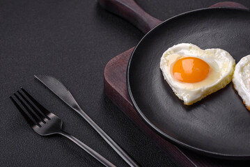 Two heart-shaped fried eggs on a black ceramic plate on a dark concrete background