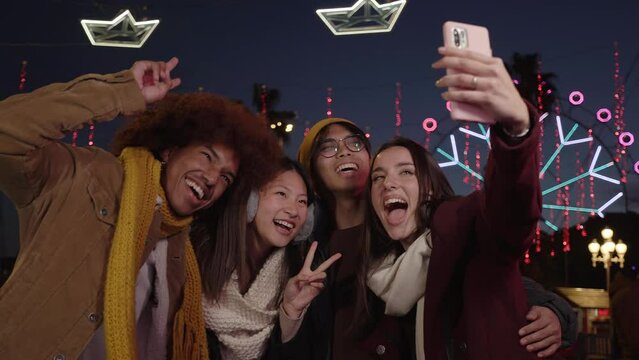Young girl and her group of friends making a video call from the amusement park. Four happy colleagues looking at their cell phones standing together at night. Multi ethnic group posing outdoor.