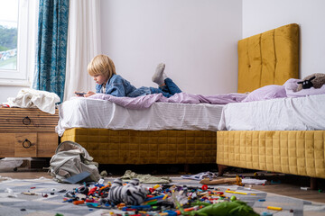 boy using phone, playing phone games in messy childrens room, kid playing among the many toys at...