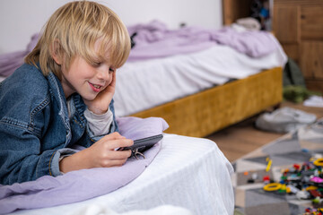 boy using phone, playing phone games in messy childrens room, kid playing among the many toys at...