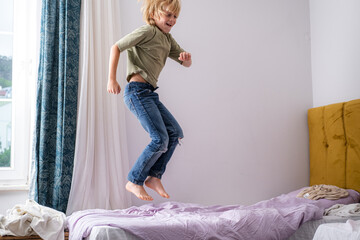 boy having fun, jumping on bed in messy childrens room, kid playing among the many toys at home, a...
