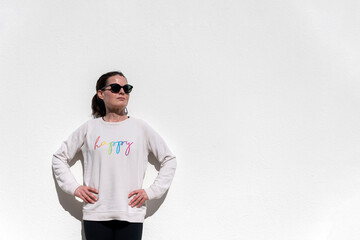 Woman standing against a white wall with hands on hips wearing a sweatshirt with happy slogan on it.
