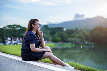 Happy Relaxing Portrait asian woman Wear dark blue dress while sitting on green grass lawn beside a reservoir at the city park outdoors.