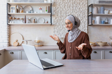 Joyful woman in hijab working from home remotely, Muslim woman talking on video call using headset and laptop, businesswoman working remotely at home.