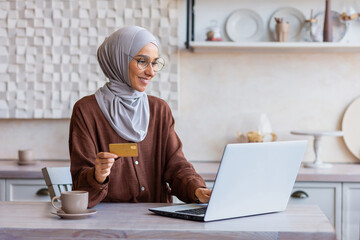 Cheerful and successful woman in hijab at home in kitchen with laptop and bank credit card makes online purchase in online store, Muslim woman chooses products and gifts remotely.