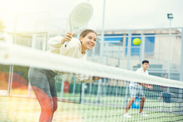 Cheerful fit young girl paddle tennis player waiting to receive serve, ready to perform forehand to return ball to opponent field on outdoor court on summer day
