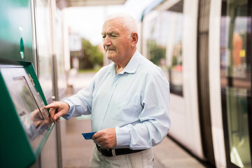 Elderly Caucasian man using cash machine to withdraw his money while standing on tram station.