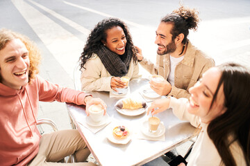Group of young multiracial people having breakfast in an outdoor coffee shop. Students of different countries having fun together drinking coffees and cappuccino sitting on a cafeteria table. High 