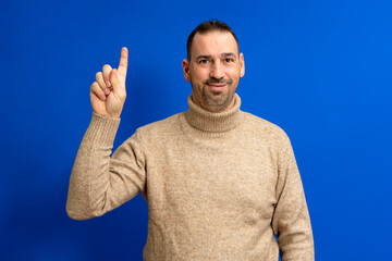 Naklejka premium Insightful intelligent proactive man hispanic man 40s wearing turtleneck holding index finger up with great new idea isolated on blue background studio portrait. People lifestyle concept.