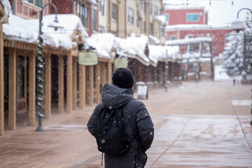 Fototapeta premium Man stands in snow wind's mist. On the building roofs, there is a thick layer of snow. In order to keep drivers and passengers safe from slick conditions and accidents, snow is cleared from the street