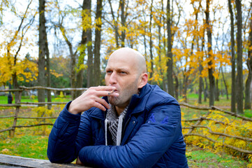 bald young man in a jacket smokes a cigarette on a bench in the autumn park. a man enjoys a calm...
