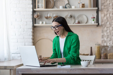 A young woman is sitting at home in the kitchen using a laptop. Plays online games, lottery, raffle. Celebrates victory, rejoices in winning