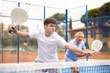 Young guy playing doubles paddle tennis with mature male partner on outdoors court, active...