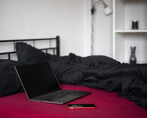 Laptop with black blank screen and smartphone on bed with viva magenta bed sheet, black blanket and pillows in modern apartment. Freelancer, blogger, business outsourcing. Copy space, mockup, template