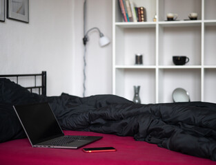 Laptop with black blank screen and smartphone on bed with viva magenta bed sheet, black blanket and pillows in modern apartment. Freelancer, blogger, business outsourcing. Copy space, mockup, template