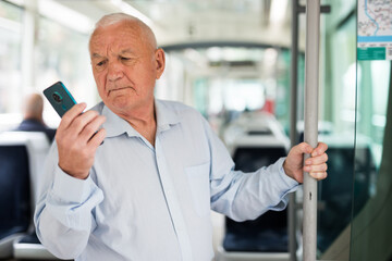 Senior European man with smartphone in hand standing inside tram and waiting for his stop.