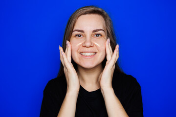 Cute young blonde woman with natural smooth skin without makeup posing happy and smiling at camera while standing in t-shirt on blue background. Beauty concept, eye patches for women