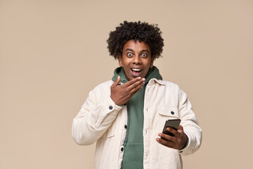 Excited surprised African teen guy holding cell phone receiving message with wow promotion. Amazed ethnic teenager student boy winner using mobile feeling shocked isolated on beige background. - obrazy, fototapety, plakaty