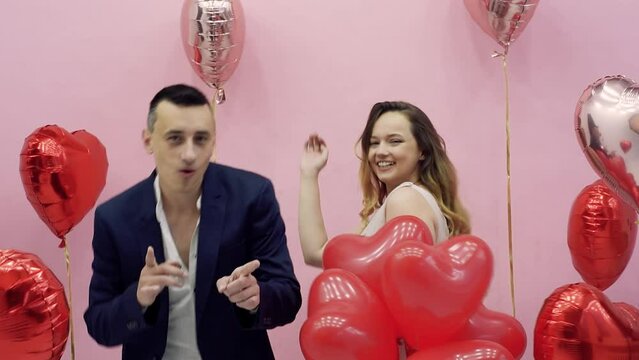 Beautiful Valentine's Day couple in red and pink balloons celebrating February 14, love. A woman in an evening dress and a guy in a suit are dancing, laughing and having fun.