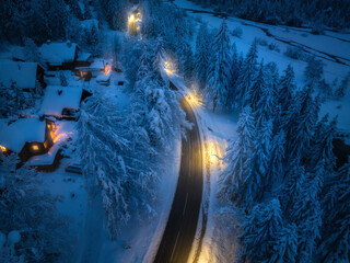 Aerial view of road in fairy town in snow, forest, houses with street lights at night in winter. Top view from drone of village, rural road, illumination, snowy pine trees at dusk in Slovenia 