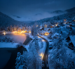 Aerial view of fairy town in snow, road, forest, Jasna lake and houses with lights at night in winter. Top view from drone of mountain village, pond, illumination, snowy pine trees at dusk in Slovenia