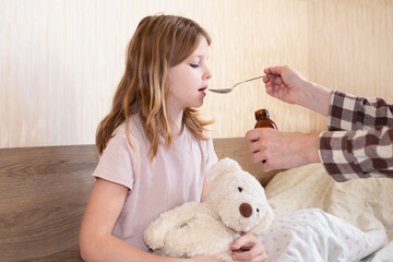 parents give medicine to child. parents at home give medicine to a cute sick girl in a spoon. sick girl hugging a teddy bear
