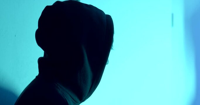 Silhouette of a guy in a hood against the background of a flashing light. The hacker is watching the work. No face, covert surveillance.