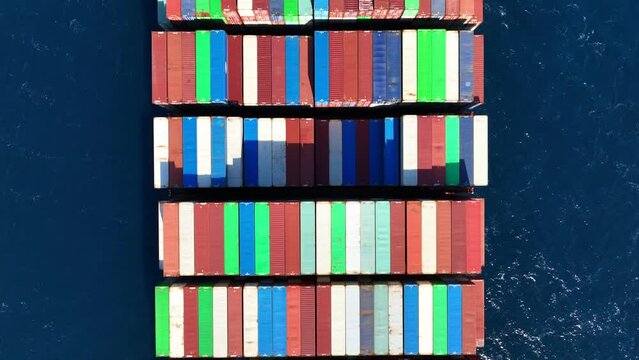Aerial drone top down tracking video of huge Container cargo Ship carrying load in truck-size colourful containers cruising deep blue Aegean sea, Greece