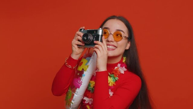 Happy young woman tourist photographer in sunglasses taking pictures photos on retro camera. Travel, summer holiday vacation, trip to seaside. Lovely smiling girl traveler on studio red background