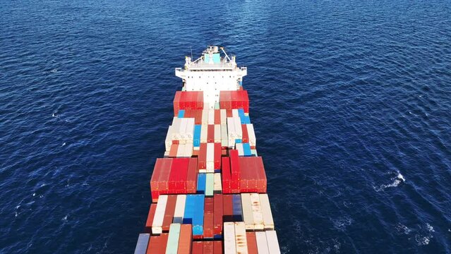 Aerial drone top down tracking video of huge Container cargo Ship carrying load in truck-size colourful containers cruising deep blue Aegean sea, Greece