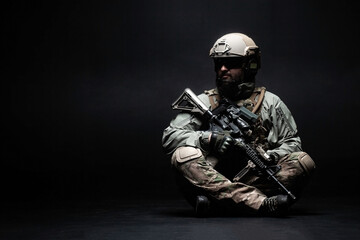 American soldier in a military uniform with a weapon sitting against a dark background and looking at the copy space, elite troops, American special forces