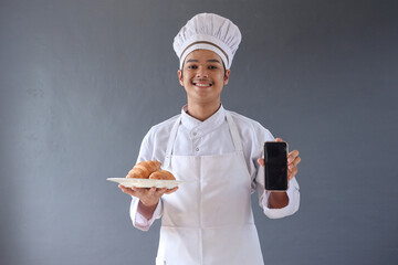 Young male chef or baker man in apron hold a plate of croissant while showing blank smartphone screen