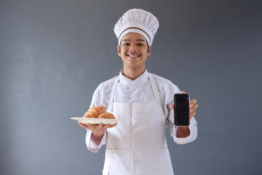 Young male chef or baker man in apron hold a plate of croissant while showing blank smartphone screen
