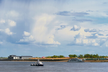 Fototapeta na wymiar Clouds over the Amur River with ships on a summer day. Border boat anchored in the middle of the river and pleasure boat on the pier. Green trees and buildings on the Chinese coast. View from Russia