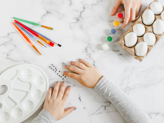 Children's hands, eggs in a box, paints, brushes and a palette on a marble table.