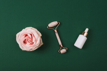 White cosmetic bottles, facial roller and pink rose flower on dark green background. Natural cosmetics, skin care concept. Top view, flat lay