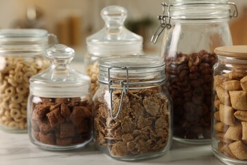 Glass containers with different breakfast cereals on white marble table indoors