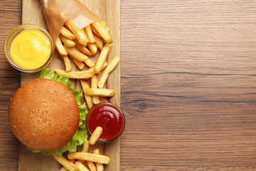 French fries, tasty burger and sauces on wooden table, top view. Space for text
