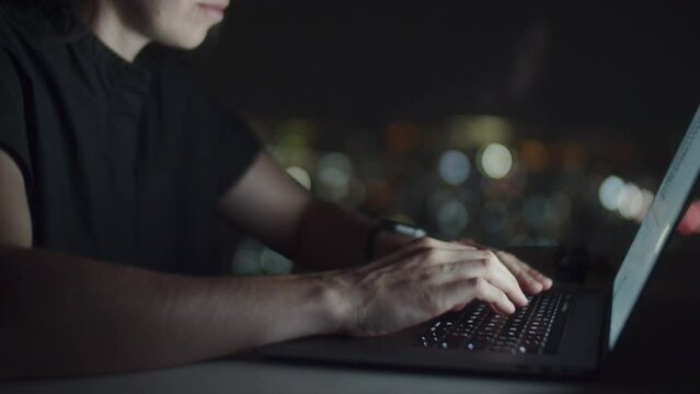 Woman typing computer, sitting at desk late night with white glowing screen and lights on background. Work during non-working hours