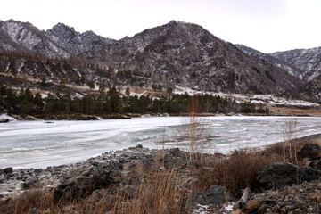 The frozen bed of a wide river flowing through a picturesque valley at the foot of snow-covered mountain ranges.