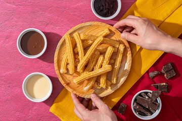 Crop person serving plate with churros, top view of crop anonymous person putting wooden plate with fresh sweet churros on pink table near bowls with various toppings.